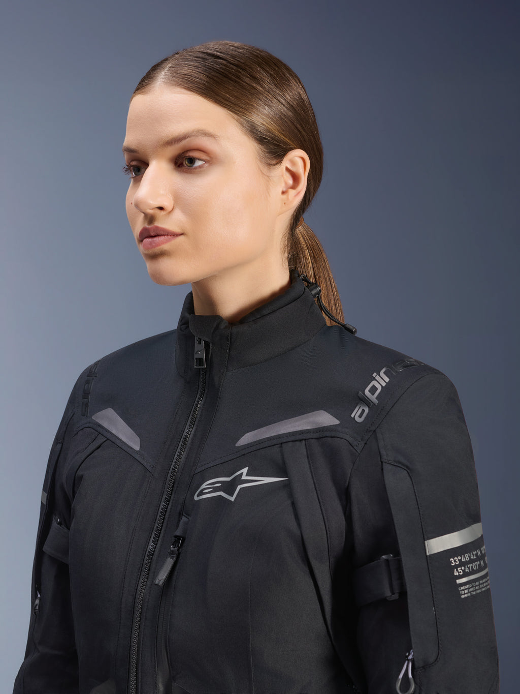 Alpinestars Stella ST-7 2L GORE-TEX damesmotorjas, zwart, voorzien van logo op de borst en technische coördinaten op de arm, waterdicht textielontwerp met hoge kraag voor touring, studioportret van een vrouwelijke rijder.