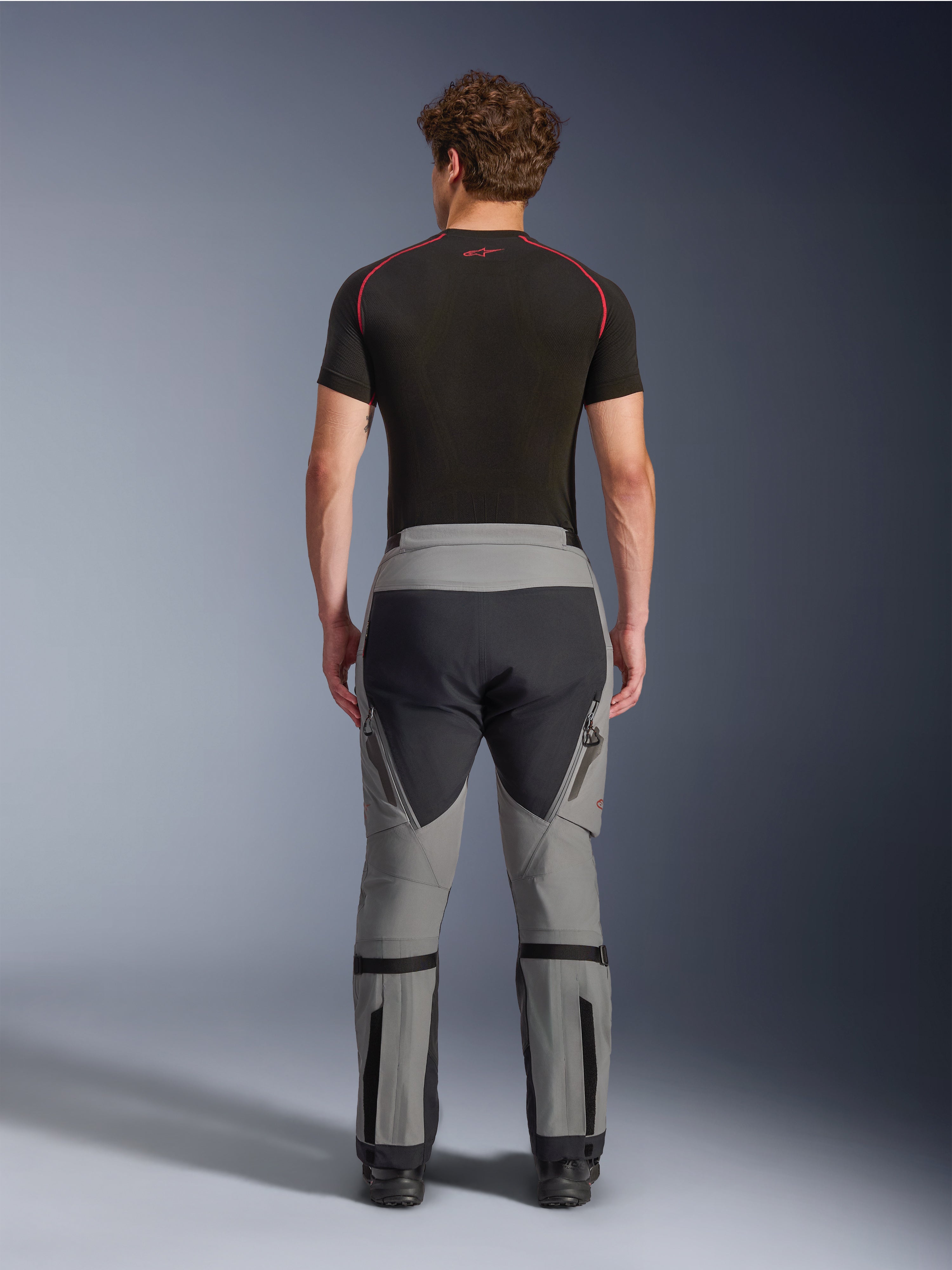 Alpinestars Nazca 3L GORE-TEX PRO-broek, Dark Gray Black adventure-motorbroek, achteraanzicht op een model, technisch multi-panel ontwerp met grijze en zwarte stof, voorzien van kuitvolume-verstellers en gedragen met een zwarte basislaag met rode accenten.