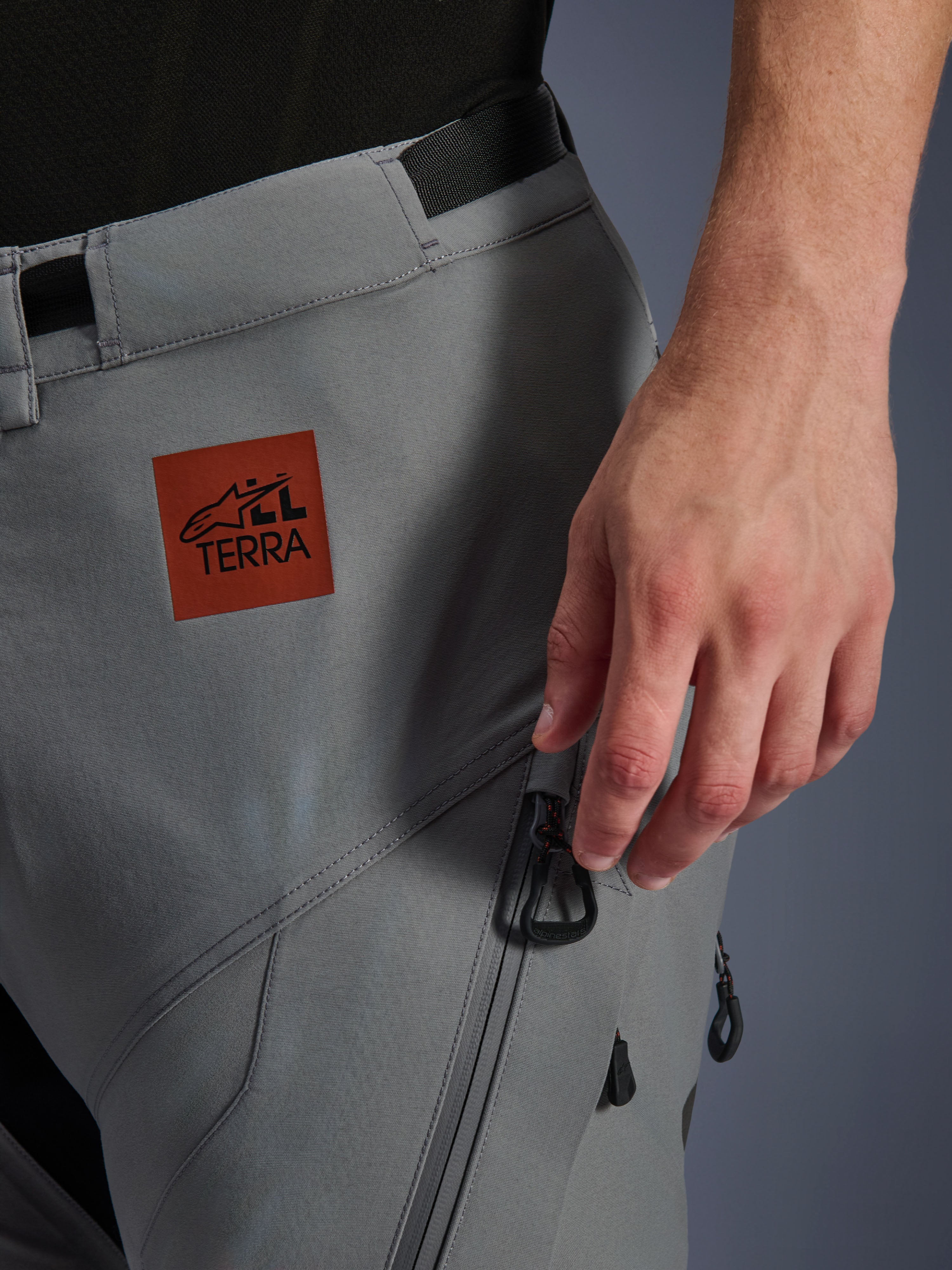 Alpinestars Nazca 3L GORE-TEX PRO broek, adventure motorbroek, donkergrijs, close-up van het bovenbeen met een roodbruine All Terra logo patch, cargozak met ritssluiting en zwarte trekkers, duurzame textielconstructie voor rijden op elk terrein