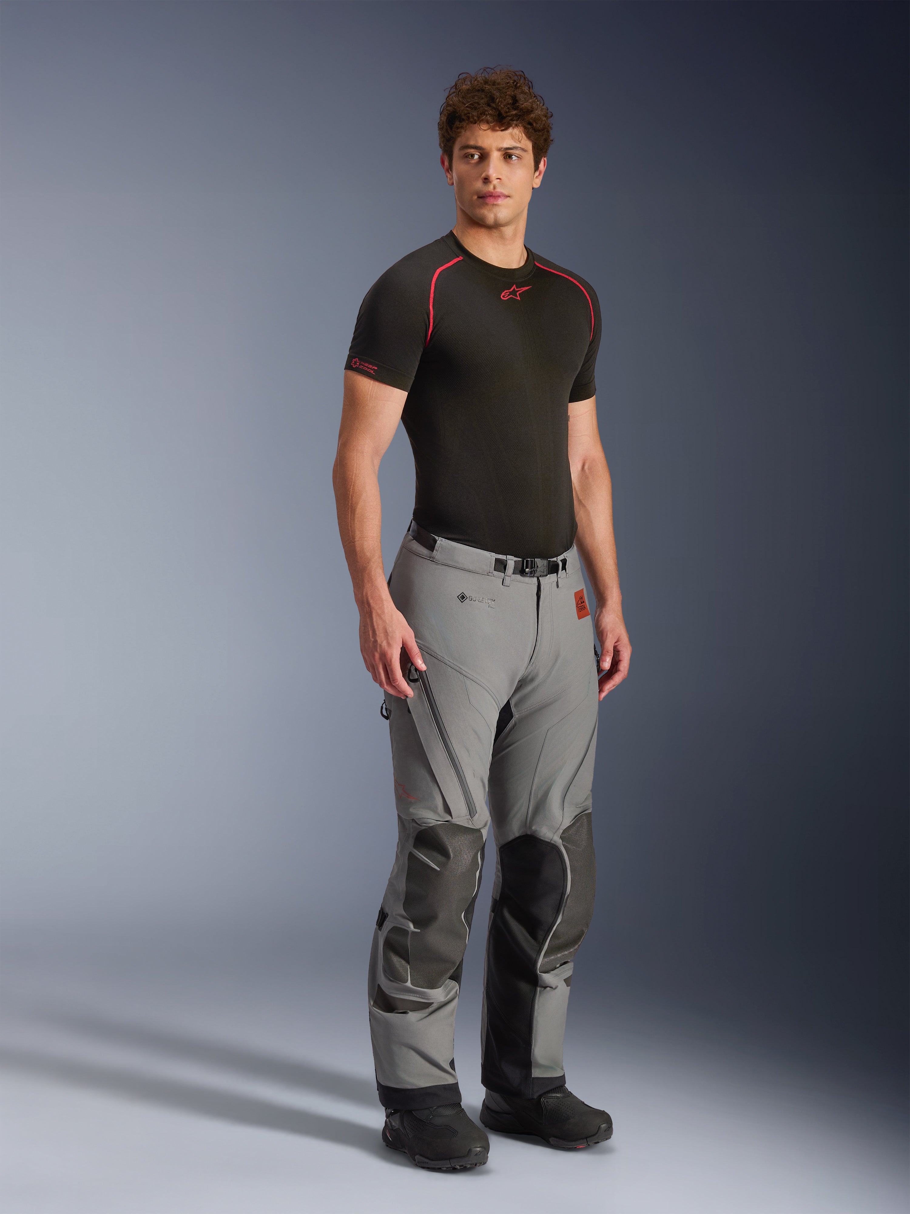 Mannelijk model draagt Alpinestars Nazca 3L GORE-TEX PRO broek, donkergrijze en zwarte adventure motorbroek, voorzien van versterkte kniepanelen en ventilatie met ritssluiting, gedragen met een zwarte base layer top, technische uitrusting ontworpen voor extreem all-terrain rijden.