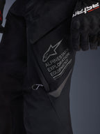 Close-up van de Alpinestars ST-7 2L GORE-TEX broek in het zwart, gedetailleerde weergave van de grote cargozak op de dij met grijze Alpinestars Exploration Equipment branding, sterlogo en geografische coördinaten, hoogwaardige textielconstructie voor adventure touring.