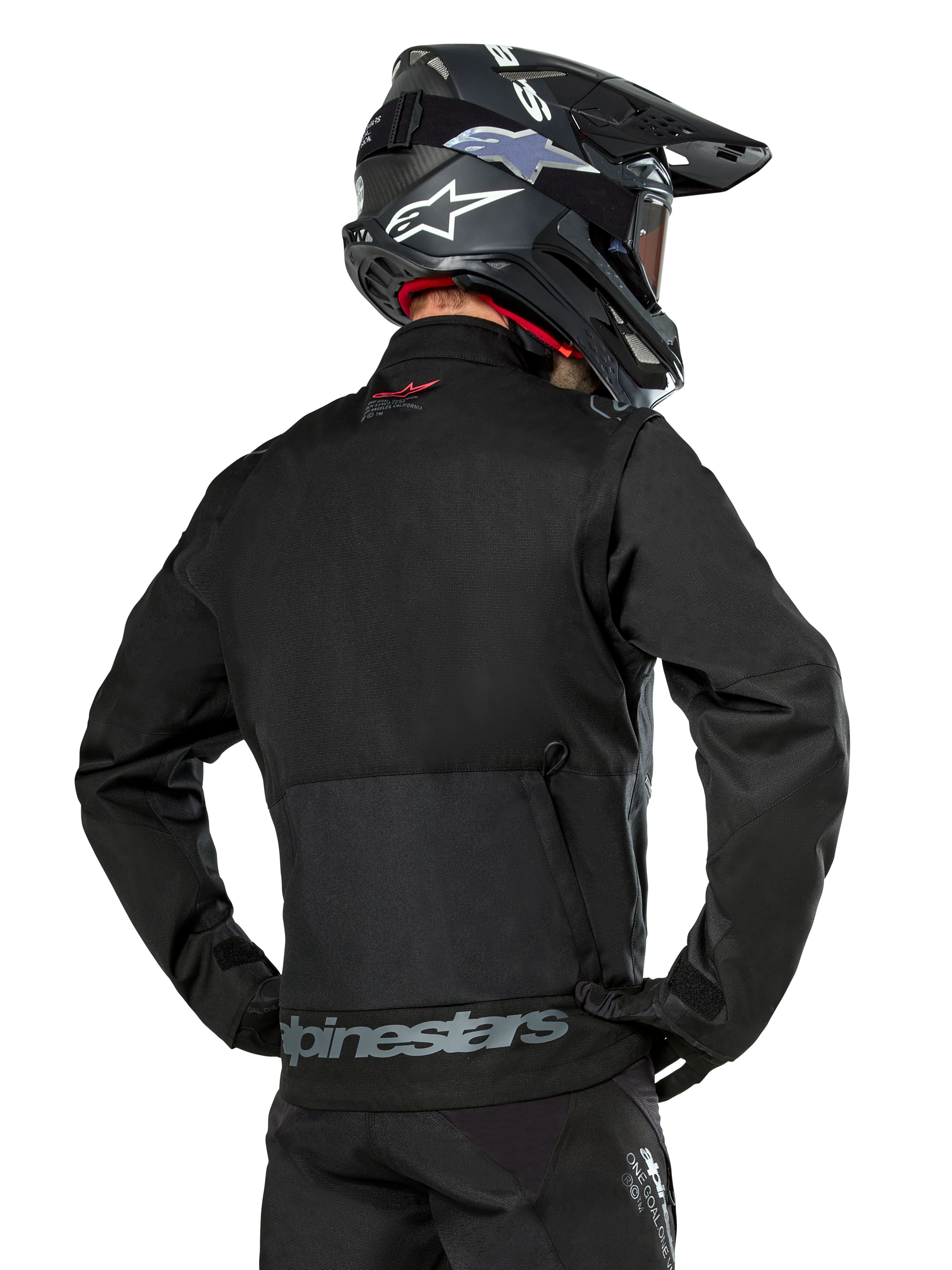Back view of a rider wearing the Alpinestars Pro-Dura Jacket in Black, durable ripstop textile enduro jacket, featuring a large rear cargo pocket with gray Alpinestars branding across the lower back, paired with a black motocross helmet.