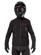 Motorcrosser met Alpinestars Lite-Dura Softshell Jas in het zwart, voorzien van rode en grijze logo-details op de borst, zwarte helm met rood getinte bril, zwarte handschoenen en broek, vooraanzicht van technische off-road motorkleding
