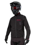 Alpinestars Lite-Dura Softshell Jacket, zwarte off-road motorjas met rode branding en AS-logo, gedragen met een zwarte motorcrosshelm, rode motorbril en zwarte handschoenen, voorzien van handzakken met ritssluiting en verstelbare manchetten, ontworpen voor enduro-rijden.