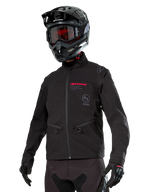 Alpinestars Lite-Dura Softshell Jacket, zwarte off-road motorjas met rode branding en AS-logo, gedragen met een zwarte motorcrosshelm, rode motorbril en zwarte handschoenen, voorzien van handzakken met ritssluiting en verstelbare manchetten, ontworpen voor enduro-rijden.