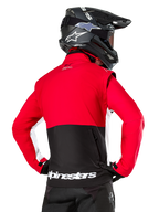 Alpinestars Lite-Dura Softshell Jacket, Black Bright Red White, achteraanzicht op een motorcrosser, rood bovenstuk en mouwen met een zwart paneel op de onderrug, opbergzak met rits op de rug, wit Alpinestars-logo op de onderste zoom, gedragen met een zwarte helm en broek