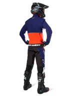 Motorcrosser vanaf de achterkant in Alpinestars Lite-Dura Softshell Jas in navy, feloranje en wit, met grote opbergzak op de rug, gecombineerd met bijpassende endurobroek, zwarte motorcrosshelm en zwarte off-road laarzen