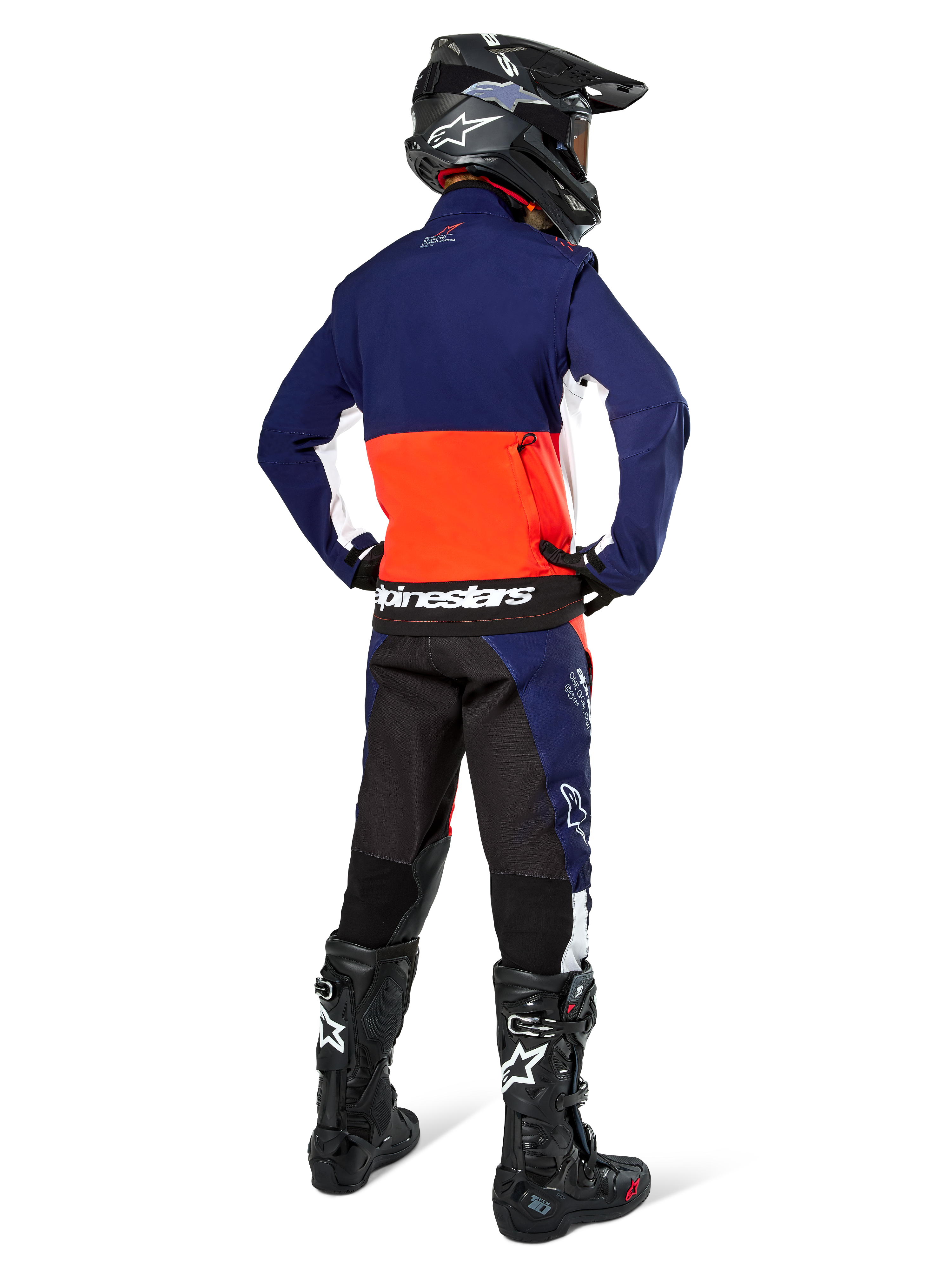Motorcrosser vanaf de achterkant in Alpinestars Lite-Dura Softshell Jas in navy, feloranje en wit, met grote opbergzak op de rug, gecombineerd met bijpassende endurobroek, zwarte motorcrosshelm en zwarte off-road laarzen