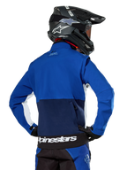 Alpinestars Lite-Dura Softshell Jas in navy, felblauw en wit, achteraanzicht met de grote achterzak met rits voor het opbergen van de mouwen, witte zij-accenten en Alpinestars-branding op de zoom, gedragen met een zwarte motorcrosshelm en bijpassende uitrusting