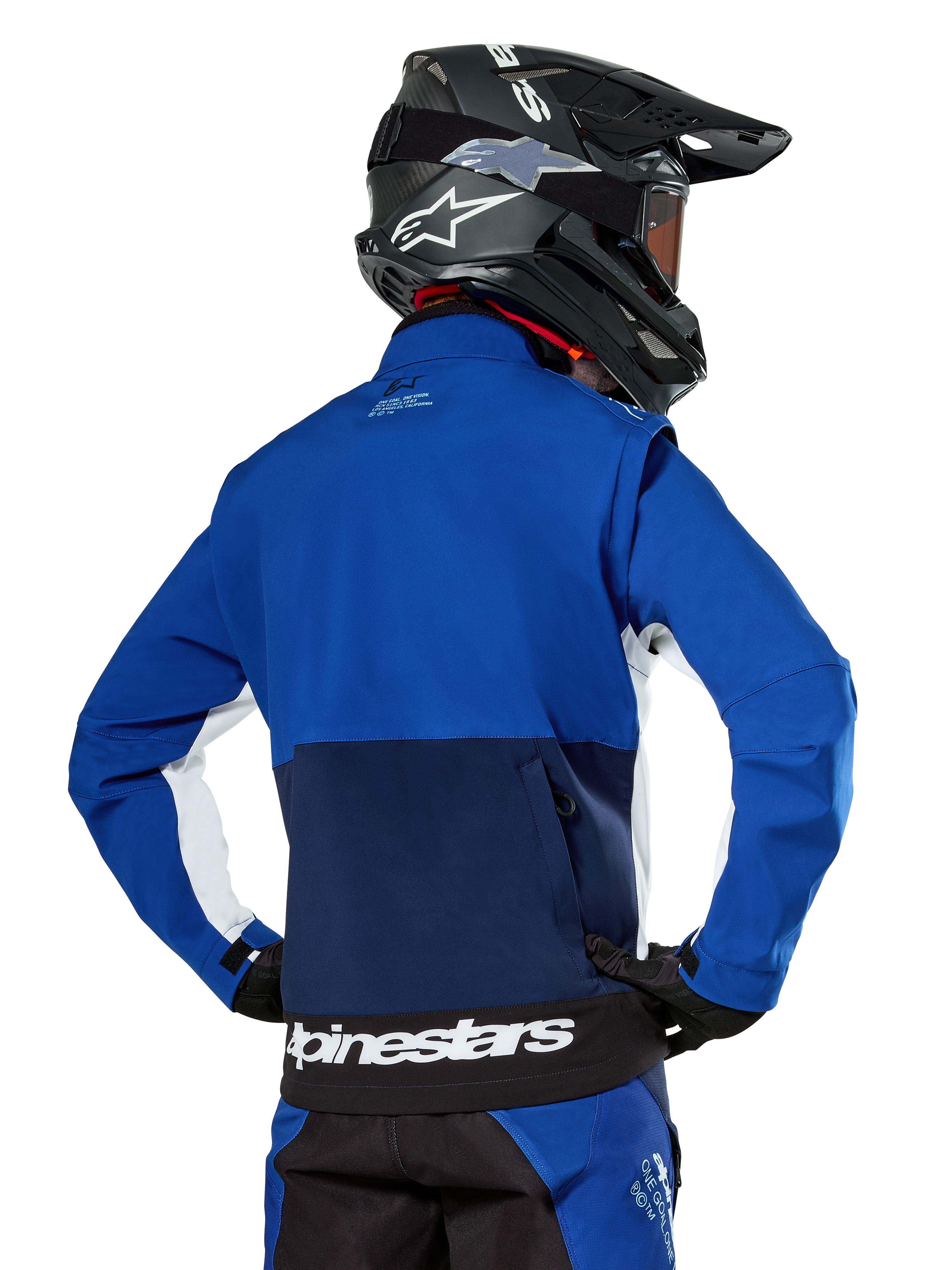 Alpinestars Lite-Dura Softshell Jas in navy, felblauw en wit, achteraanzicht met de grote achterzak met rits voor het opbergen van de mouwen, witte zij-accenten en Alpinestars-branding op de zoom, gedragen met een zwarte motorcrosshelm en bijpassende uitrusting
