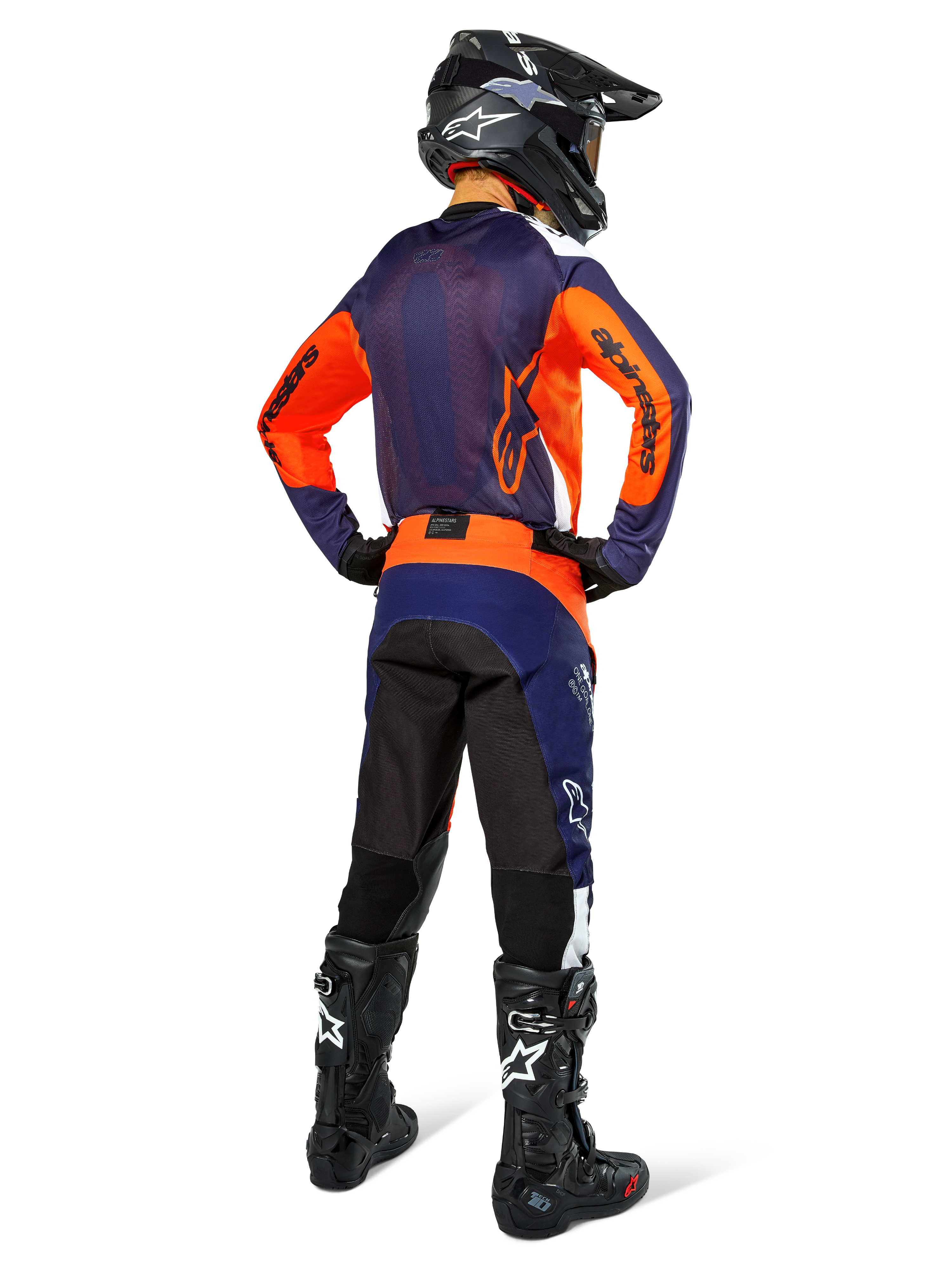Alpinestars motocrossrijder van achteren gezien met volledige race-uitrusting, marineblauwe en oranje jersey, Pro-Dura broek in Navy Hot Orange White, zwarte helm en zwarte laarzen, professionele off-road race-outfit ensemble.