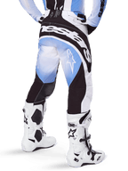 Alpinestars 2025 Techstar Melt crossbroek, motorcrossbroek, zwart, blauw en wit, achteraanzicht met opvallend Alpinestars-woordmerk op het zitvlak en sterlogo's op de pijpen, gedragen met witte en zwarte Tech 10-laarzen, professionele off-road race-uitrusting