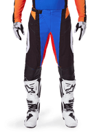 Alpinestars 2025 Techstar Melt crossbroek, oranje en blauw, gedragen met bijpassende jersey en witte laarzen, zwarte panelen met levendige blauwe en oranje accenten, geperforeerd voor ventilatie, atletische racepasvorm voor off-road prestaties