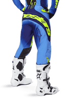 Alpinestars 2025 Techstar Melt crossbroek, motorcrossbroek, geel fluo blauw, achteraanzicht met groot geel logo op het zitvlak en donkerblauwe accenten, gedragen met witte Alpinestars Tech 10 laarzen en een bijpassende jersey.