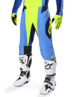 Alpinestars 2025 Techstar Melt crossbroek, motorcross broek, fluoriserend geel met blauw, gedragen met bijpassende geel-blauwe handschoenen en witte motorcrosslaarzen, met ergonomische snit en ventilatie voor off-road raceprestaties