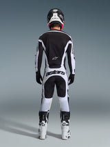 Alpinestars 2026 Racer Riway Pants, motorcrossbroek, wit en zwart, achteraanzicht van een complete race-uitrusting, opvallende 'astars' branding op het zitvlak, gedragen met bijpassend shirt, witte laarzen en zwarte helm, gecoördineerde off-road race-outfit.