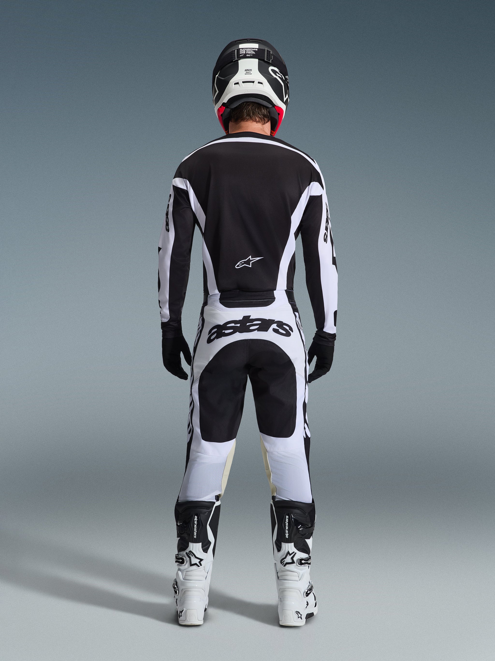 Alpinestars 2026 Racer Riway Pants, motorcrossbroek, wit en zwart, achteraanzicht van een complete race-uitrusting, opvallende 'astars' branding op het zitvlak, gedragen met bijpassend shirt, witte laarzen en zwarte helm, gecoördineerde off-road race-outfit.