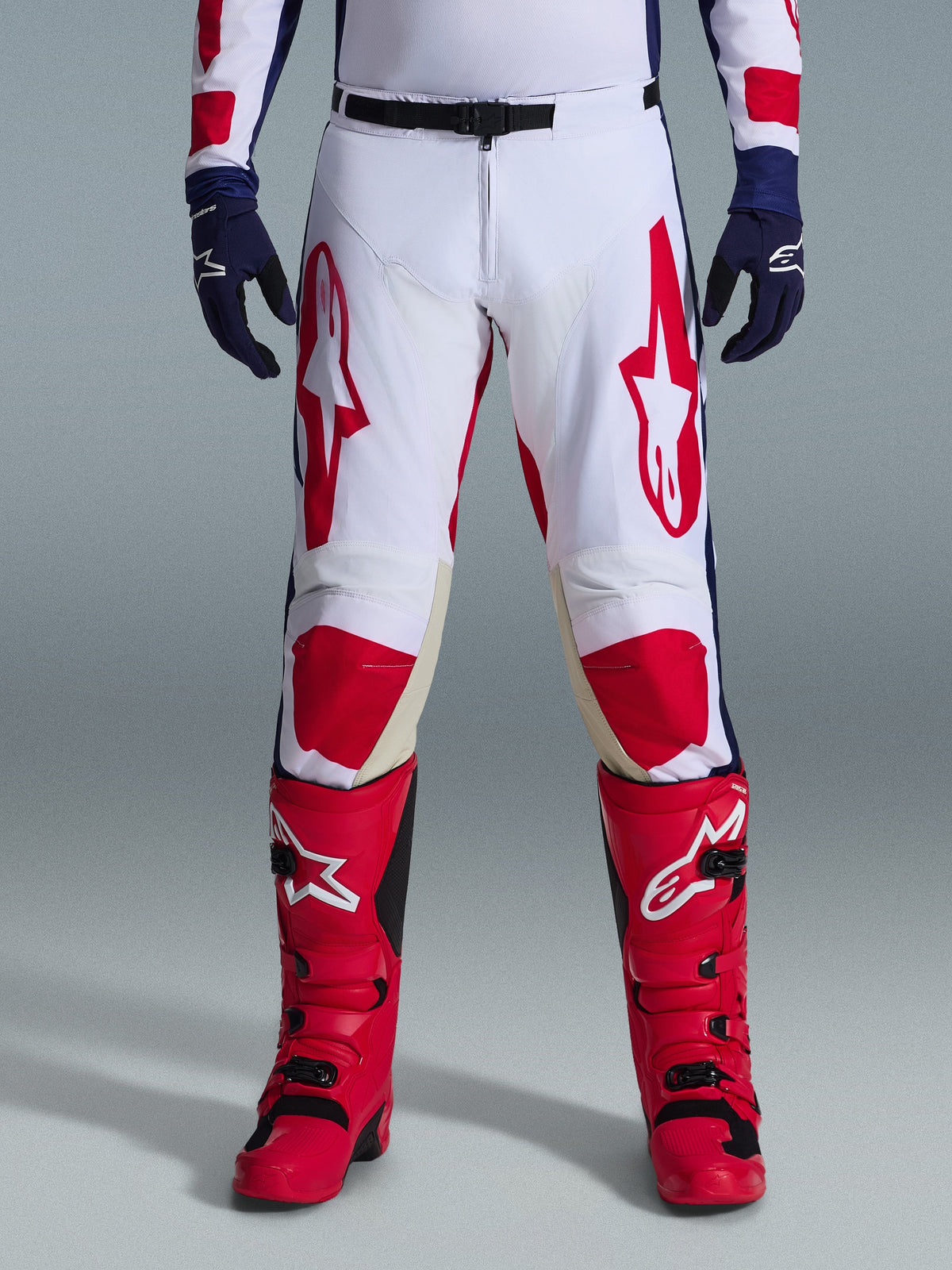 Alpinestars 2026 Racer Riway broek, motorcross broek, wit blauw en rood, gedragen met rode laarzen en donkerblauwe handschoenen, voorzien van grote rode logo's op de dijen en rode kniepanelen, sportief voorgevormd ontwerp met verstelbare gespsluiting