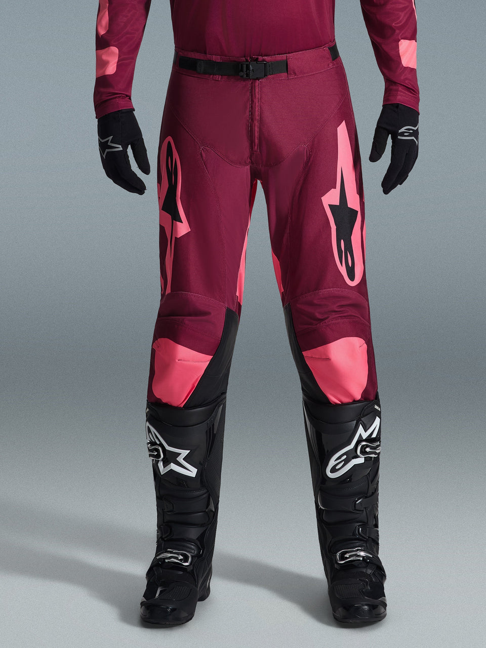 Alpinestars 2026 Racer Riway broek, motorcrossbroek, donkerrood koraal, gedragen met zwarte handschoenen en laarzen, voorzien van grote koraalkleurige Alpinestars-logo's op de dijen, zwarte gesp in de taille en voorgevormd beenontwerp voor off-road racen