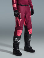 Alpinestars 2026 Racer Riway crossbroek, motorcrossbroek, Dark Red Coral, met grote roze sterlogo's en zwarte binnenpanelen op de knieën, gedragen met zwarte motorcrosslaarzen en zwarte handschoenen, atletische racepasvorm voor off-road rijden.