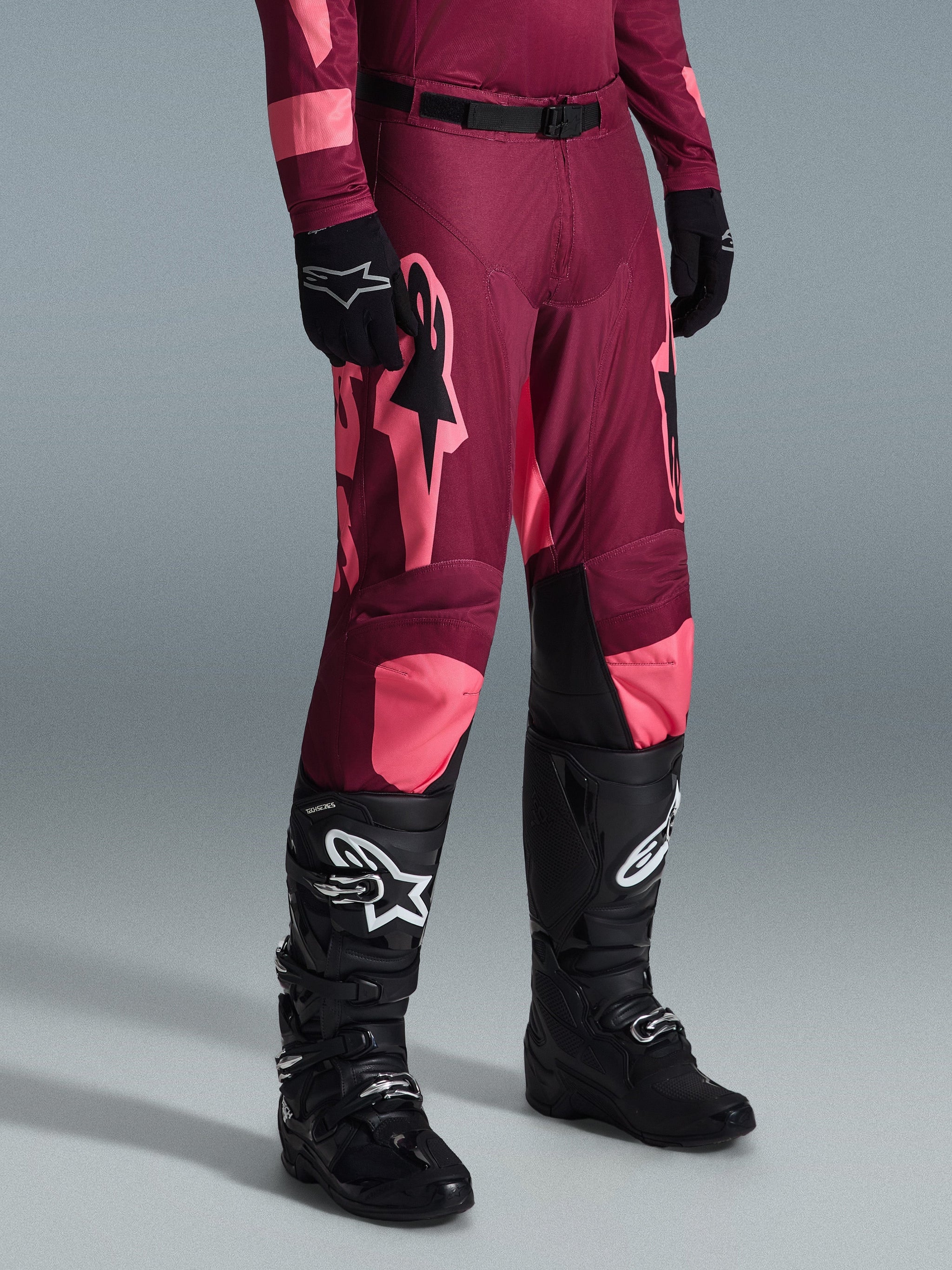 Alpinestars 2026 Racer Riway crossbroek, motorcrossbroek, Dark Red Coral, met grote roze sterlogo's en zwarte binnenpanelen op de knieën, gedragen met zwarte motorcrosslaarzen en zwarte handschoenen, atletische racepasvorm voor off-road rijden.