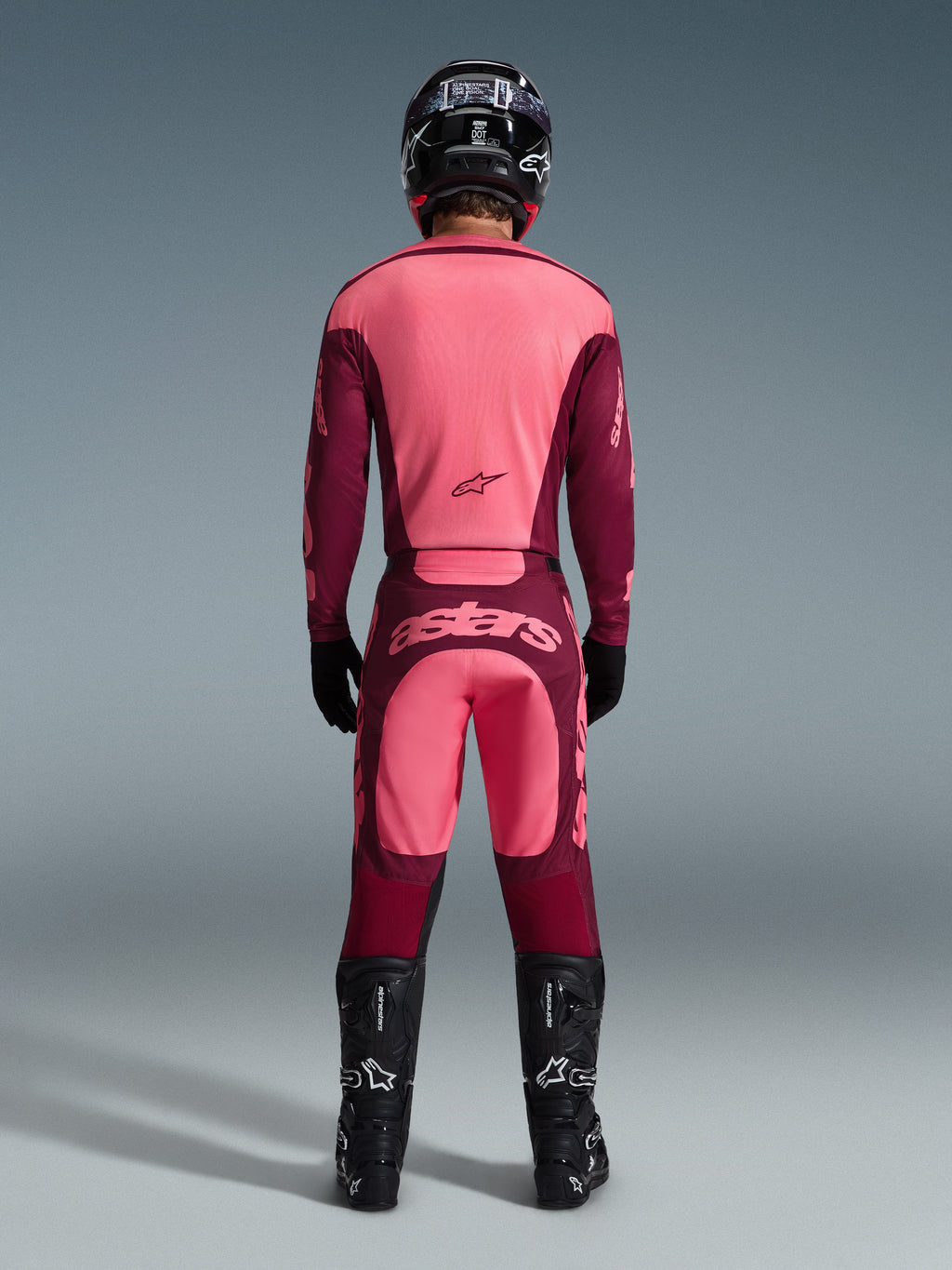 Achteraanzicht van een motorcrosser die de 2026 Racer Riway Gear Set draagt, roze en bordeauxrode jersey, bijpassende bordeauxrode broek met lichtroze 'astars'-logo op het zitvlak, zwarte helm, zwarte laarzen en zwarte handschoenen, gecoördineerde outfit voor off-road prestaties