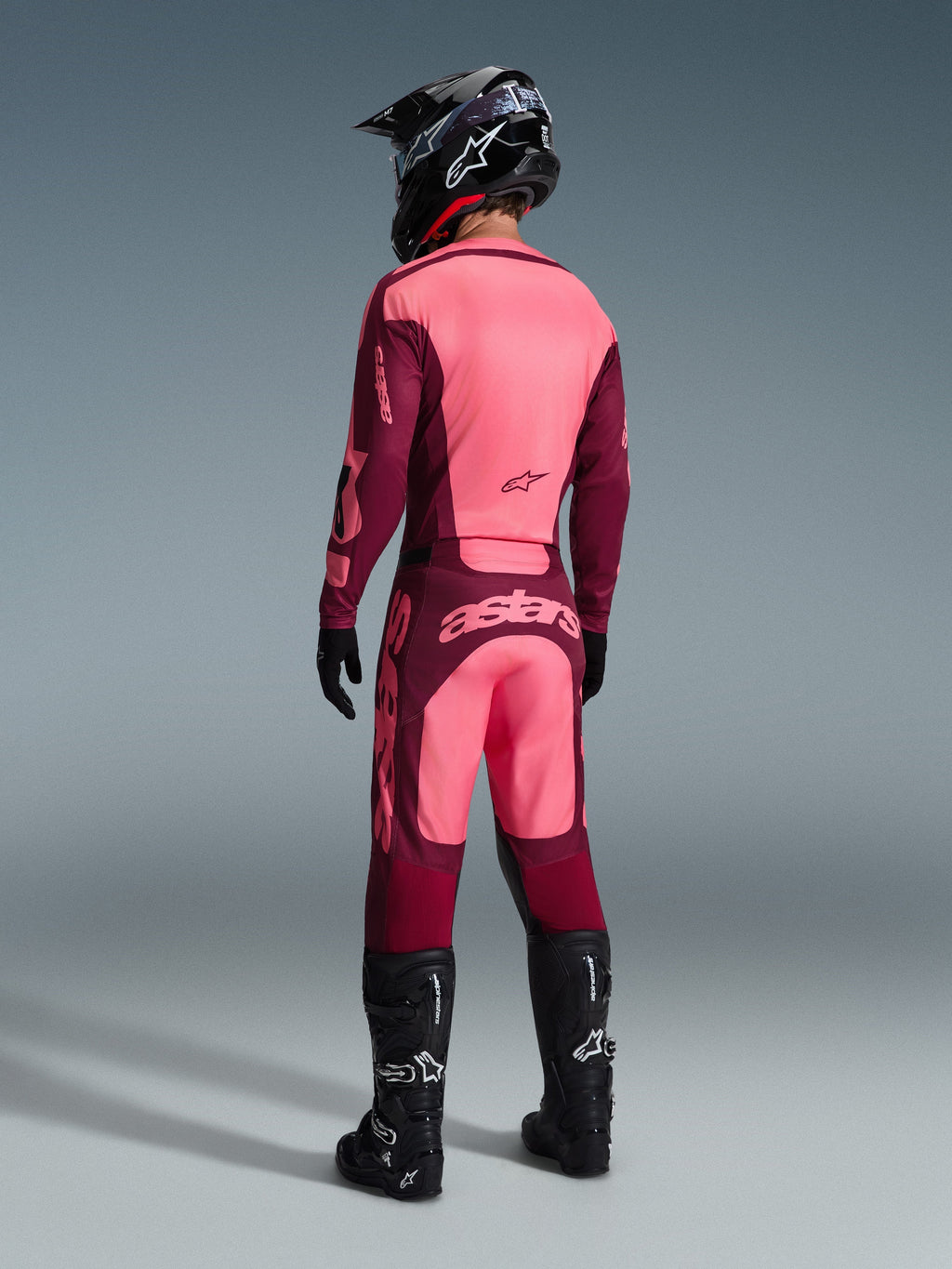Alpinestars 2026 Racer Riway broek, motorcrossbroek, Dark Red Coral, achteraanzicht van een rijder met een bijpassende jersey, zwarte helm en zwarte laarzen, met opvallende koraalrode branding op het zitvlak en een ergonomische atletische pasvorm voor off-road racen