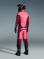 Alpinestars 2026 Racer Riway broek, motorcrossbroek, Dark Red Coral, achteraanzicht van een rijder met een bijpassende jersey, zwarte helm en zwarte laarzen, met opvallende koraalrode branding op het zitvlak en een ergonomische atletische pasvorm voor off-road racen