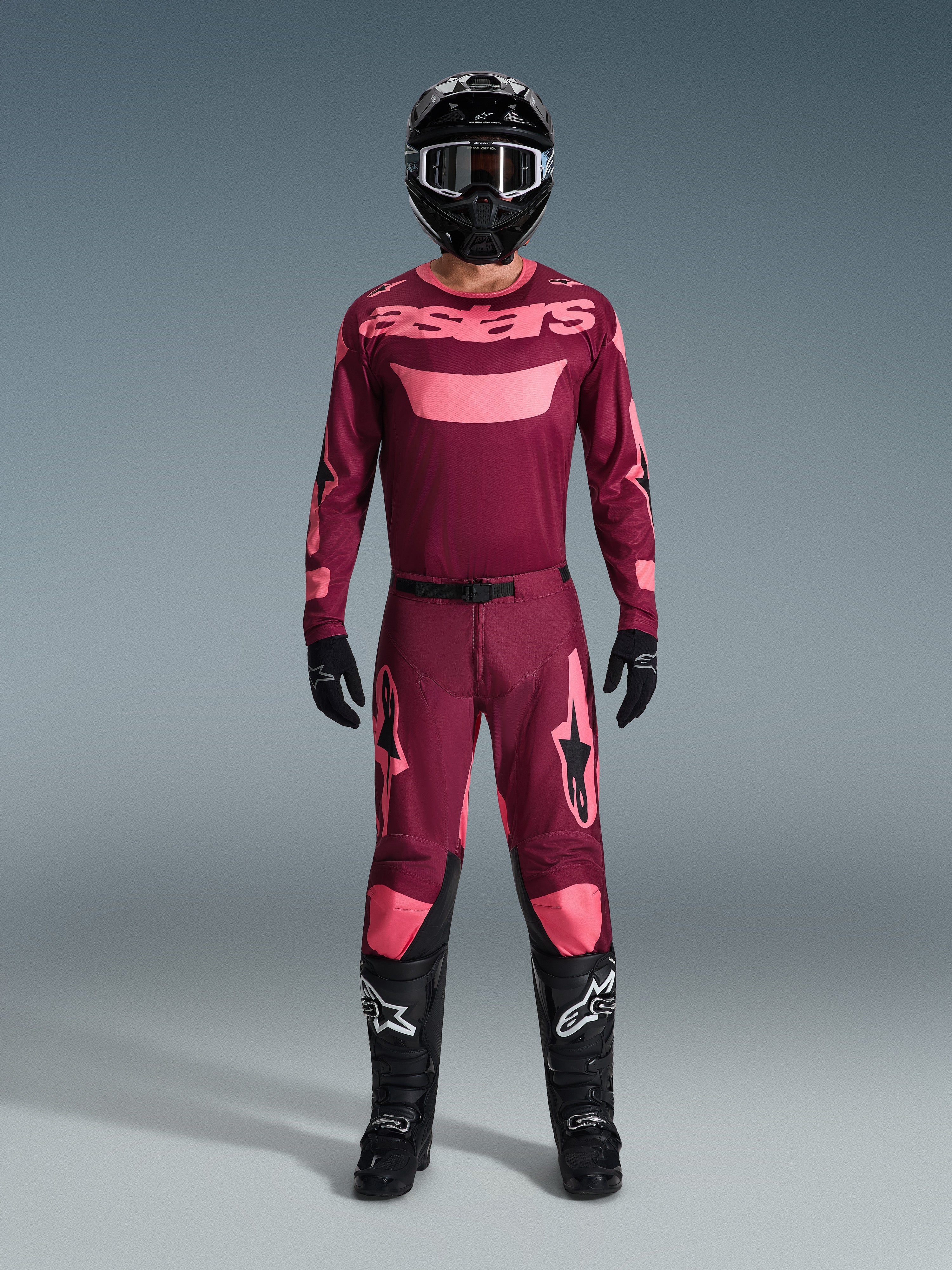 Motocross rider wearing the Alpinestars 2026 Racer Riway Gear Set, maroon jersey with pink 'astars' branding, matching maroon pants with pink knee and leg accents, black helmet, black gloves, and black boots, complete performance outfit for off-road racing
