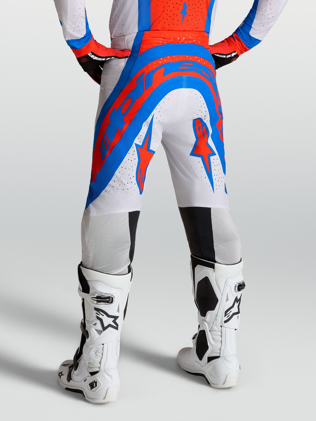 Alpinestars Supertech Vesta Broek, pro-level motocrossbroek in wit en rood met blauwe accenten, gedragen door een model met witte Tech 10 motorlaarzen, voorzien van geperforeerde stof.