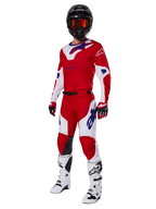 Alpinestars 2025 Racer Veil broek, motorcrossbroek, helder rood en wit, gedragen als onderdeel van een complete race-uitrusting inclusief een bijpassend rood en wit shirt, zwarte helm met oranje bril, witte laarzen en rode handschoenen, met opvallende blauwe Alpinestars-logo's.