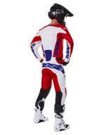 Alpinestars 2025 Racer Veil Pants, motorcrossbroek, felrood en wit met blauwe accenten, van achteren getoond op een rijder in volledige uitrusting, met opvallende Alpinestars-branding, ademende panelen op de onderbenen en bijpassende witte laarzen en een zwarte helm.