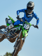 Motocrossrijder halverwege een sprong op een groene motorfiets, draagt Alpinestars 2025 Racer Veil Pants in Blauw en Zwart, bijpassende blauwe jersey met wit logo op de borst, witte en zwarte laarzen en zwarte helm, hoogwaardige off-road uitrusting voor racing.