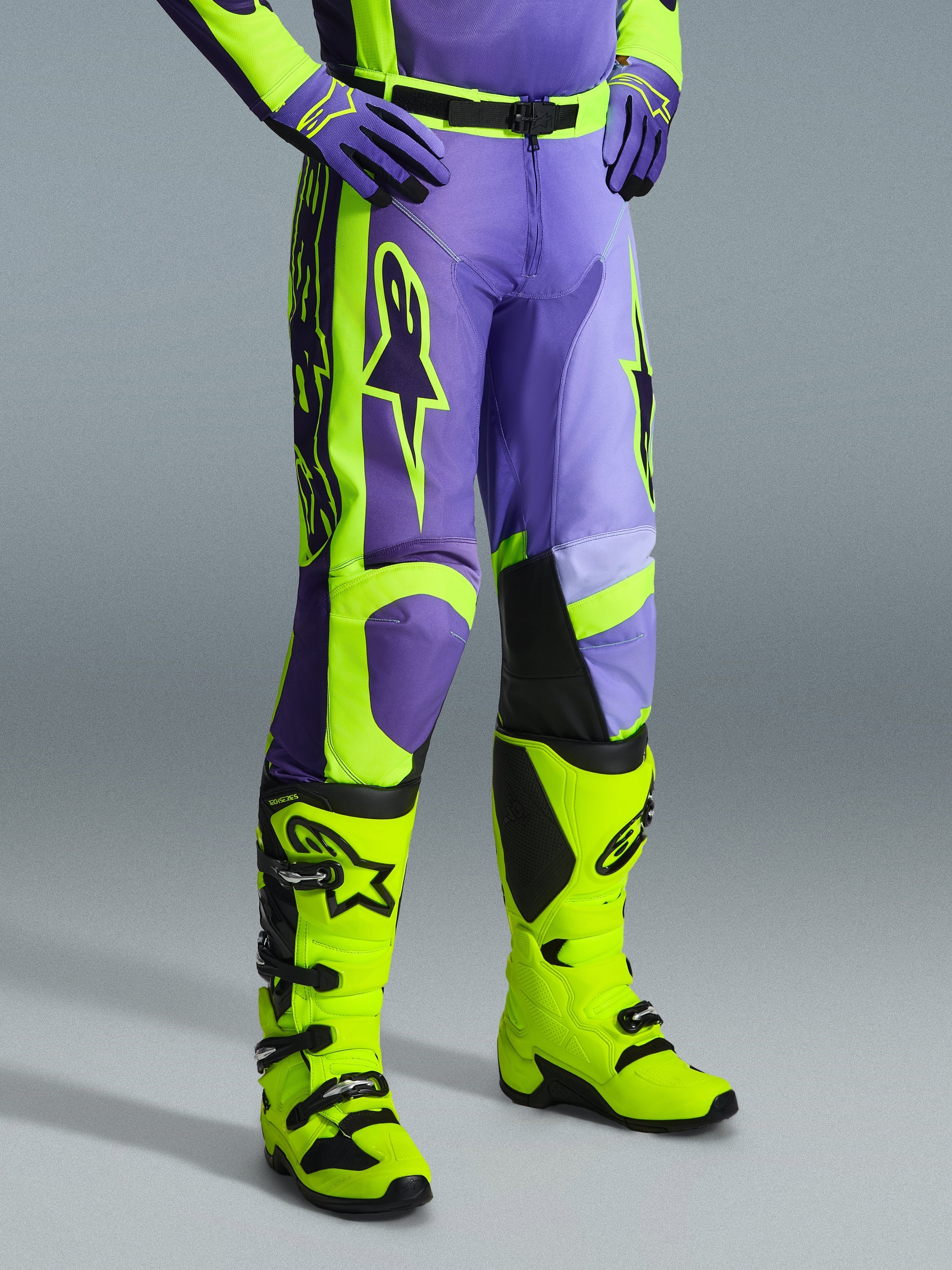 Alpinestars 2026 Racer Portl Pants, motorcrossbroek in paars en fluogeel, gedragen door een rijder met bijpassende fluogele laarzen en paarse handschoenen, met opvallende Alpinestars-branding en ergonomisch ontwerp voor off-road prestaties