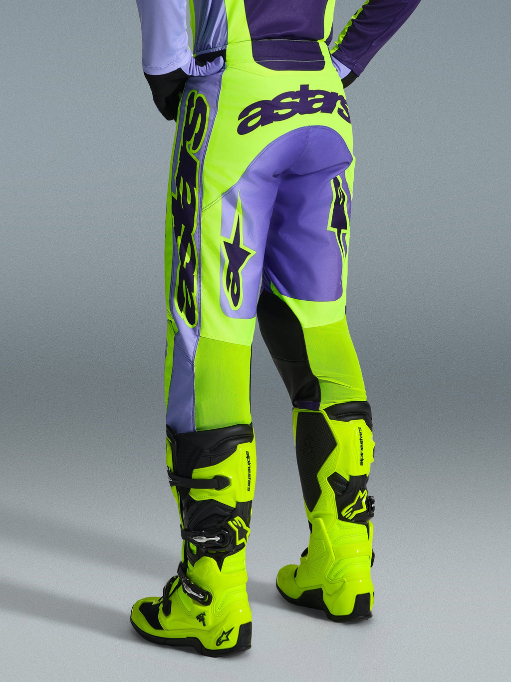 Alpinestars 2026 Racer Portl Broek, motorcrossbroek, paars en neon geel, achteraanzicht met opvallende astars-branding en paarse accenten, gedragen met bijpassende neon gele motorcrosslaarzen, duurzaam synthetisch materiaal voor off-road racing