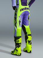 Alpinestars 2026 Racer Portl Broek, motorcrossbroek, paars en neon geel, achteraanzicht met opvallende astars-branding en paarse accenten, gedragen met bijpassende neon gele motorcrosslaarzen, duurzaam synthetisch materiaal voor off-road racing