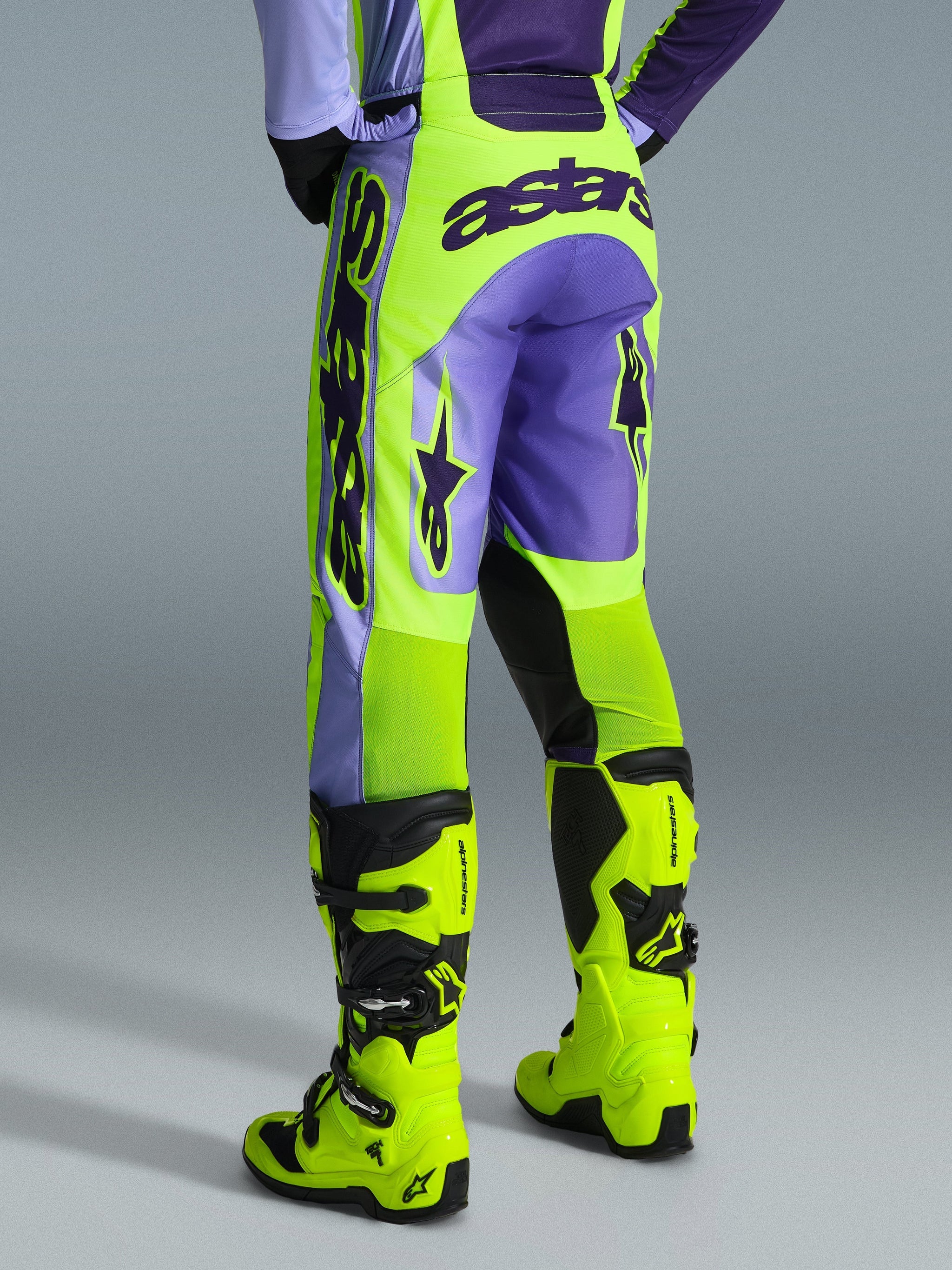 Alpinestars 2026 Racer Portl Broek, motorcrossbroek, paars en neon geel, achteraanzicht met opvallende astars-branding en paarse accenten, gedragen met bijpassende neon gele motorcrosslaarzen, duurzaam synthetisch materiaal voor off-road racing