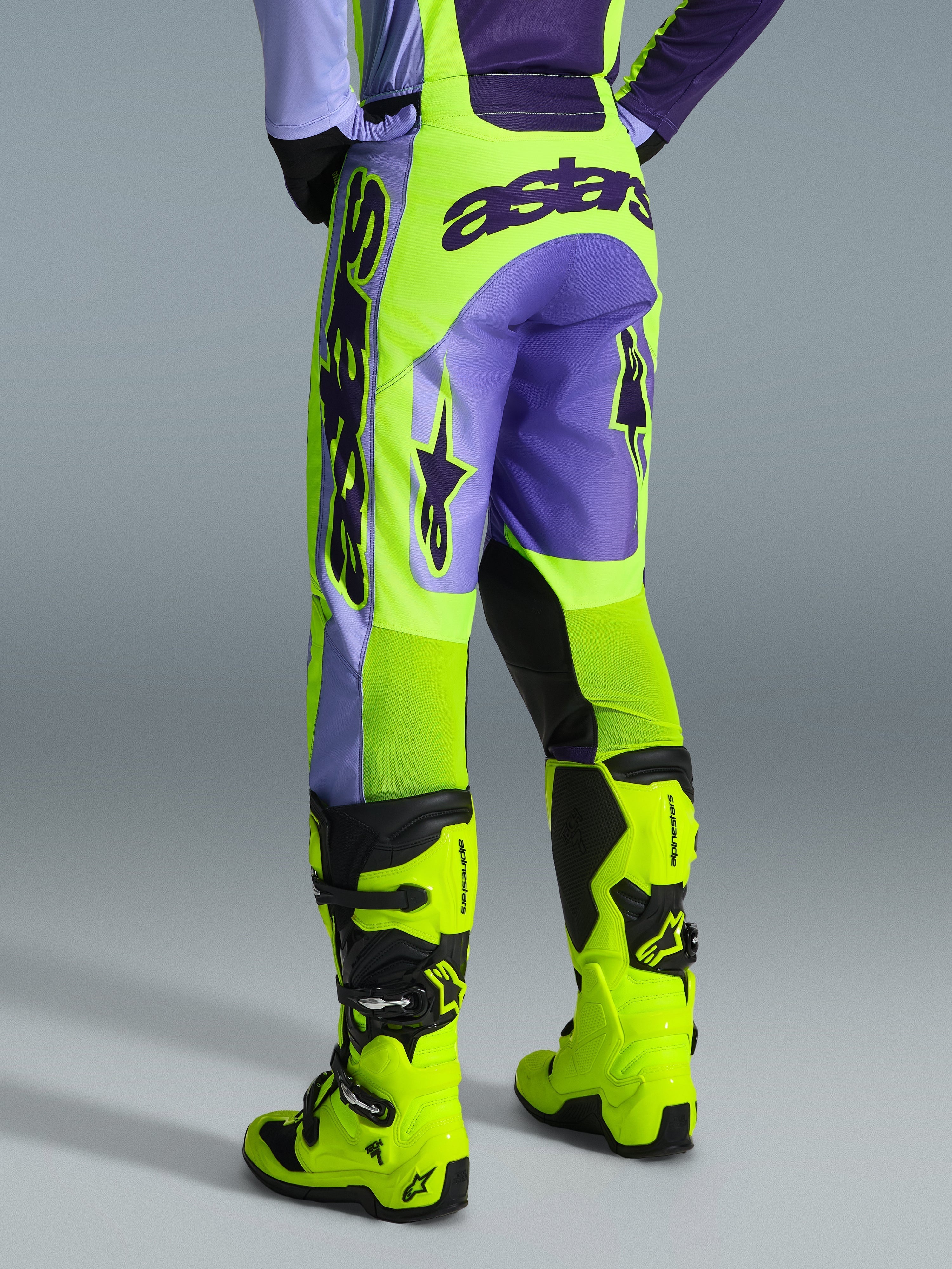 Alpinestars 2026 Racer Portl Broek, motorcrossbroek, paars en neon geel, achteraanzicht met opvallende astars-branding en paarse accenten, gedragen met bijpassende neon gele motorcrosslaarzen, duurzaam synthetisch materiaal voor off-road racing