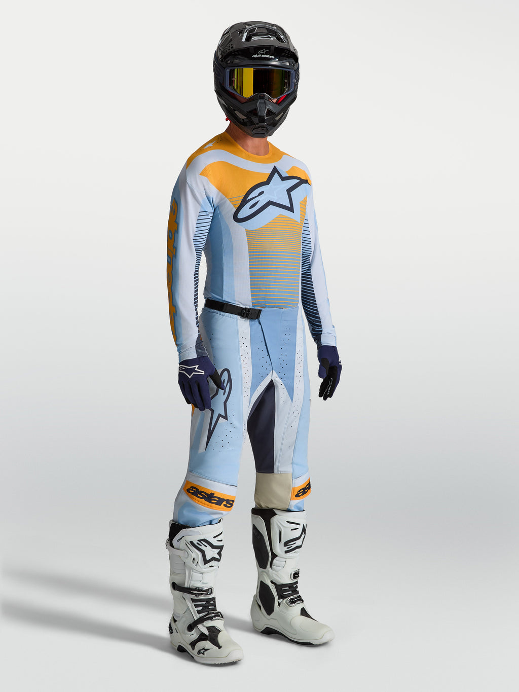 Alpinestars Techstar Maven heren-motocrossbroek, blauw en oranje, met bijpassende jersey, witte motorlaarzen en zwarte helm.