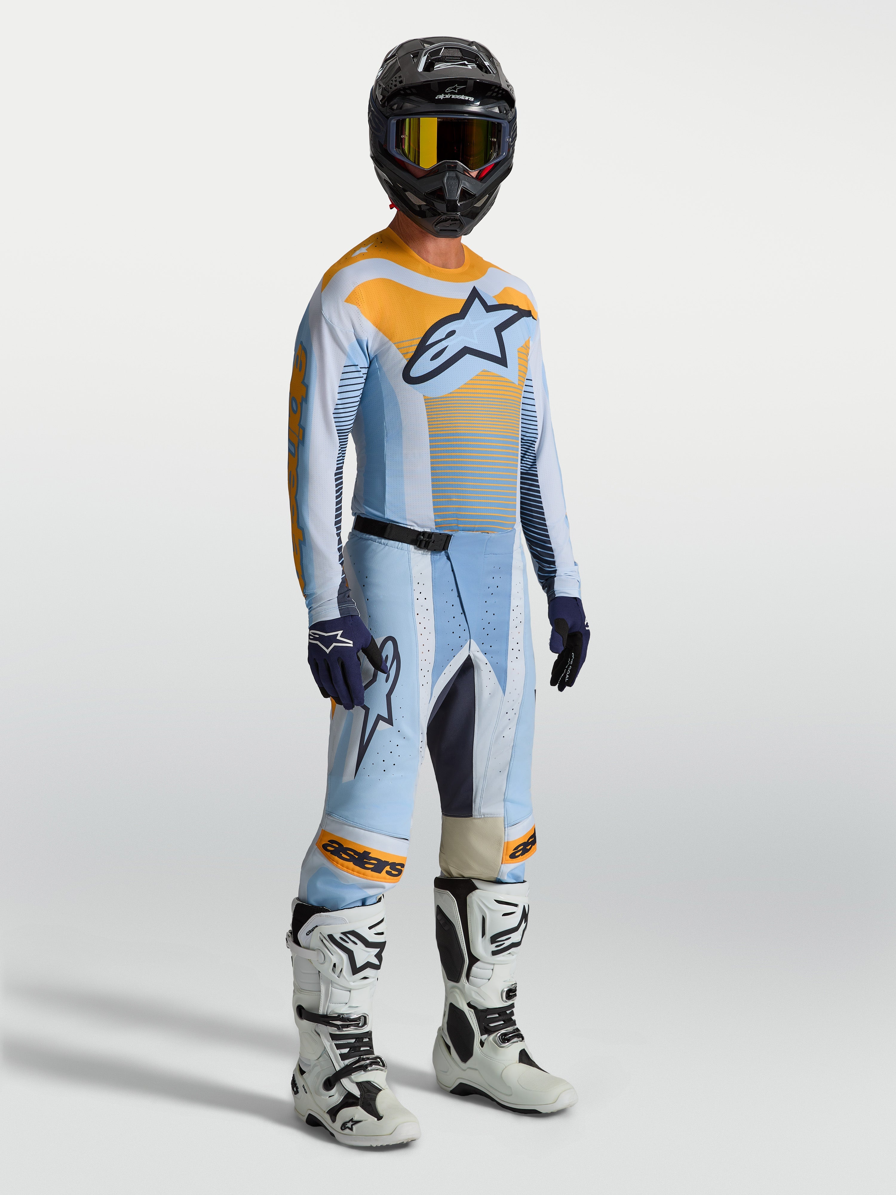 Alpinestars Techstar Maven heren-motocrossbroek, blauw en oranje, met bijpassende jersey, witte motorlaarzen en zwarte helm.