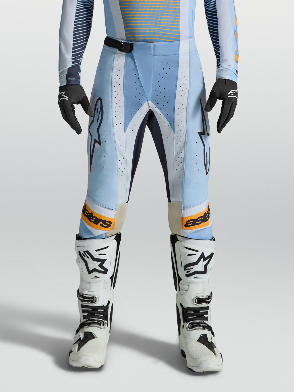 Alpinestars Techstar Maven broek, heren-motorcrossbroek in blauw en oranje, voorzien van lasergesneden ventilatie en een ergonomische racepasvorm, gedragen door een model met witte motorlaarzen.