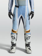 Alpinestars Techstar Maven broek, heren-motorcrossbroek in blauw en oranje, voorzien van lasergesneden ventilatie en een ergonomische racepasvorm, gedragen door een model met witte motorlaarzen.