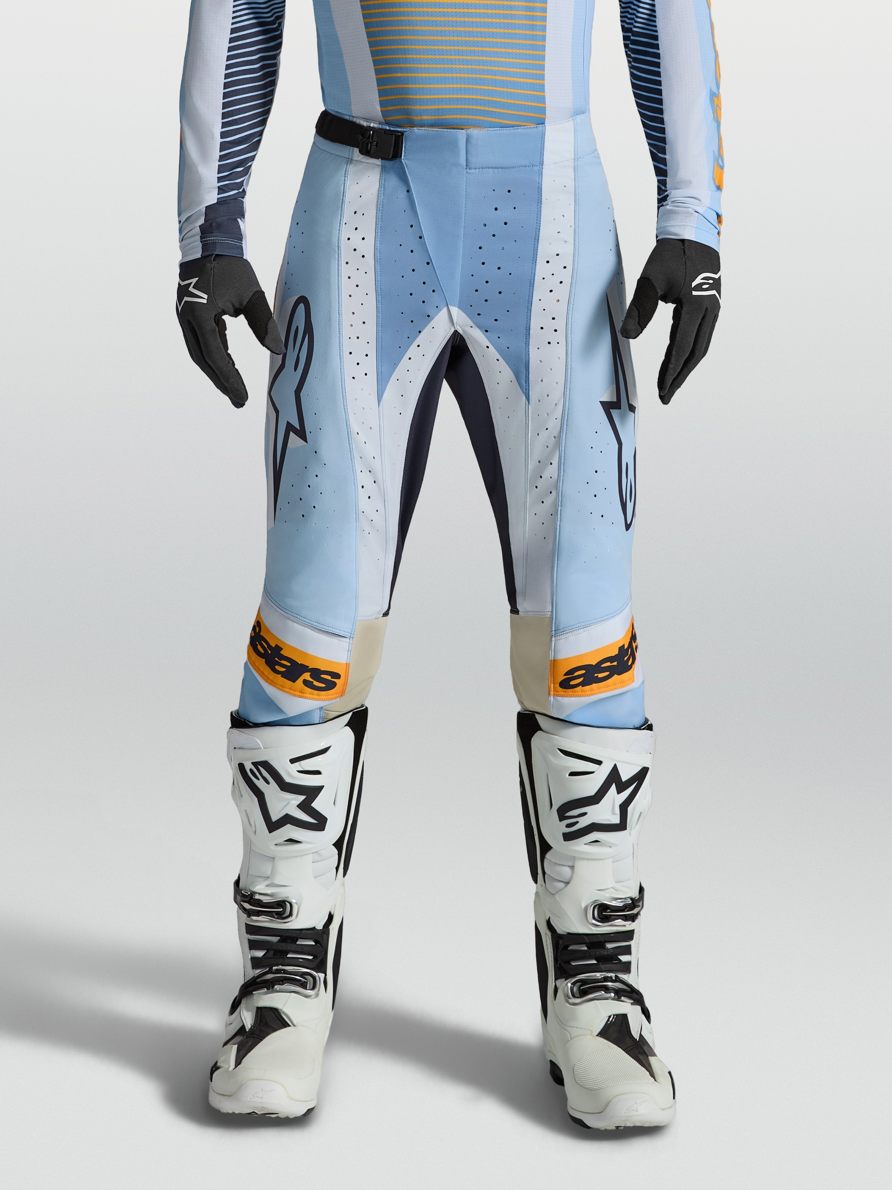 Alpinestars Techstar Maven broek, heren-motorcrossbroek in blauw en oranje, voorzien van lasergesneden ventilatie en een ergonomische racepasvorm, gedragen door een model met witte motorlaarzen.