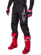 Alpinestars 2025 Fluid Haul broek, motorcrossbroek, zwart, voorzien van een groot wit Alpinestars-logo, gedragen met felrode laarzen en rood-zwarte handschoenen, duurzame 600D polyfabric constructie met ergonomische snit voor off-road racing