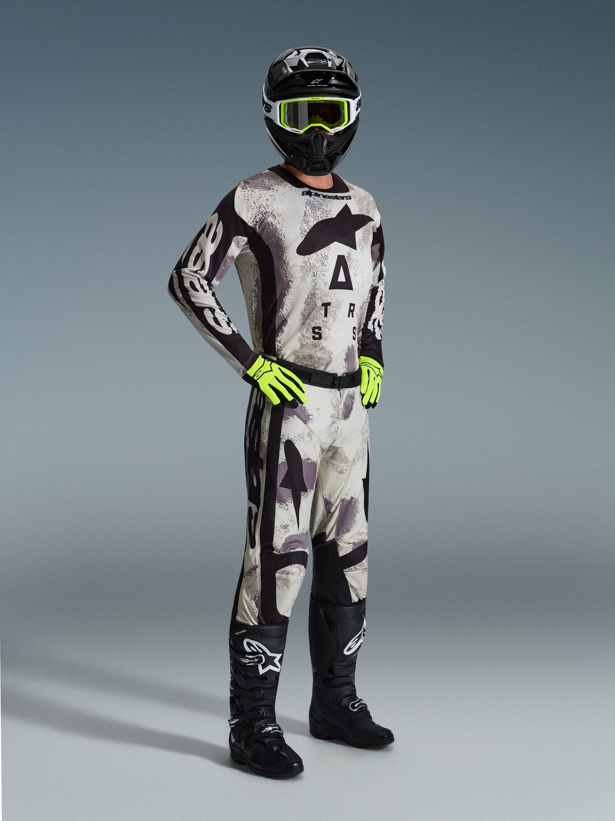Motocross-rijder in volledige Alpinestars race-uitrusting, 2026 Racer Lahnd broek in Sand Camo, bijpassend camouflage shirt met opvallende branding, zwarte helm met neongele bril, neongroene handschoenen en zwarte laarzen, gecoördineerde professionele outfit voor off-road prestaties.