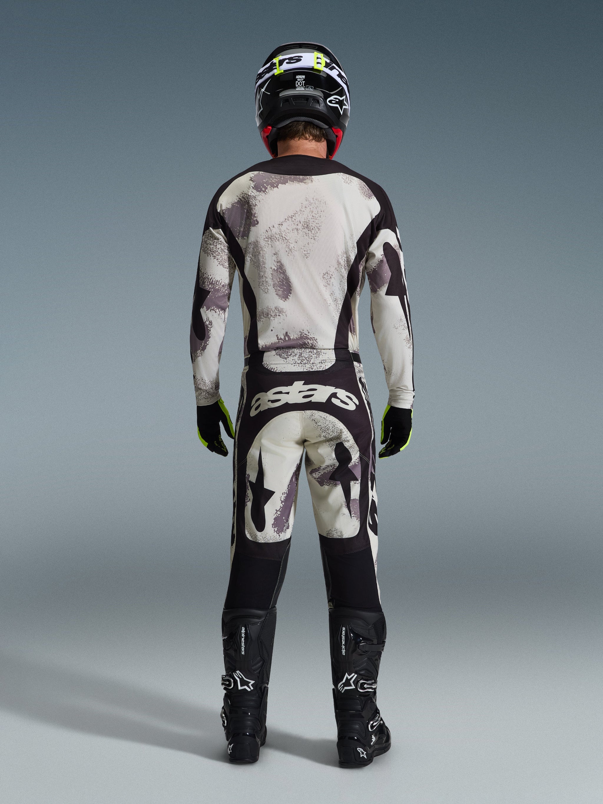 Motorcrosser van achteren in de Alpinestars 2026 Racer Lahnd broek in Sand Camo, bijpassende jersey met lange mouwen en grijs camouflagepatroon, zwarte motorcrosslaarzen, zwarte helm en 'astars' branding op het zitvlak, ontworpen voor professionele off-road racing