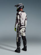 Motocross rider wearing Alpinestars 2026 Racer Lahnd Gear Set, Sand Camo abstract pattern in gray and white, black pants with bold white Alpinestars branding, black helmet and boots, fluorescent green accents on gloves, complete outfit for off-road racing