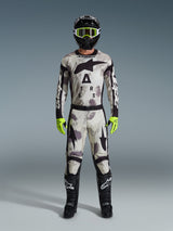 Motocrossrijder in volledige race-uitrusting, Alpinestars 2026 Racer Lahnd broek en bijpassende jersey in Sand Camo grijs, zwarte helm met neongele crossbril, felle neongele handschoenen en zwarte laarzen, gecoördineerde professionele outfit voor off-road racing