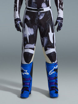 Alpinestars 2026 Racer Lahnd crossbroek, motorcross broek, Iron Camo, gedragen met bijpassende camouflage handschoenen en felblauwe en paarse laarzen, voorzien van gestileerde witte ster-graphics op een donker camouflagepatroon, technische magnetische gespsluiting, professionele off-road race-uitrusting