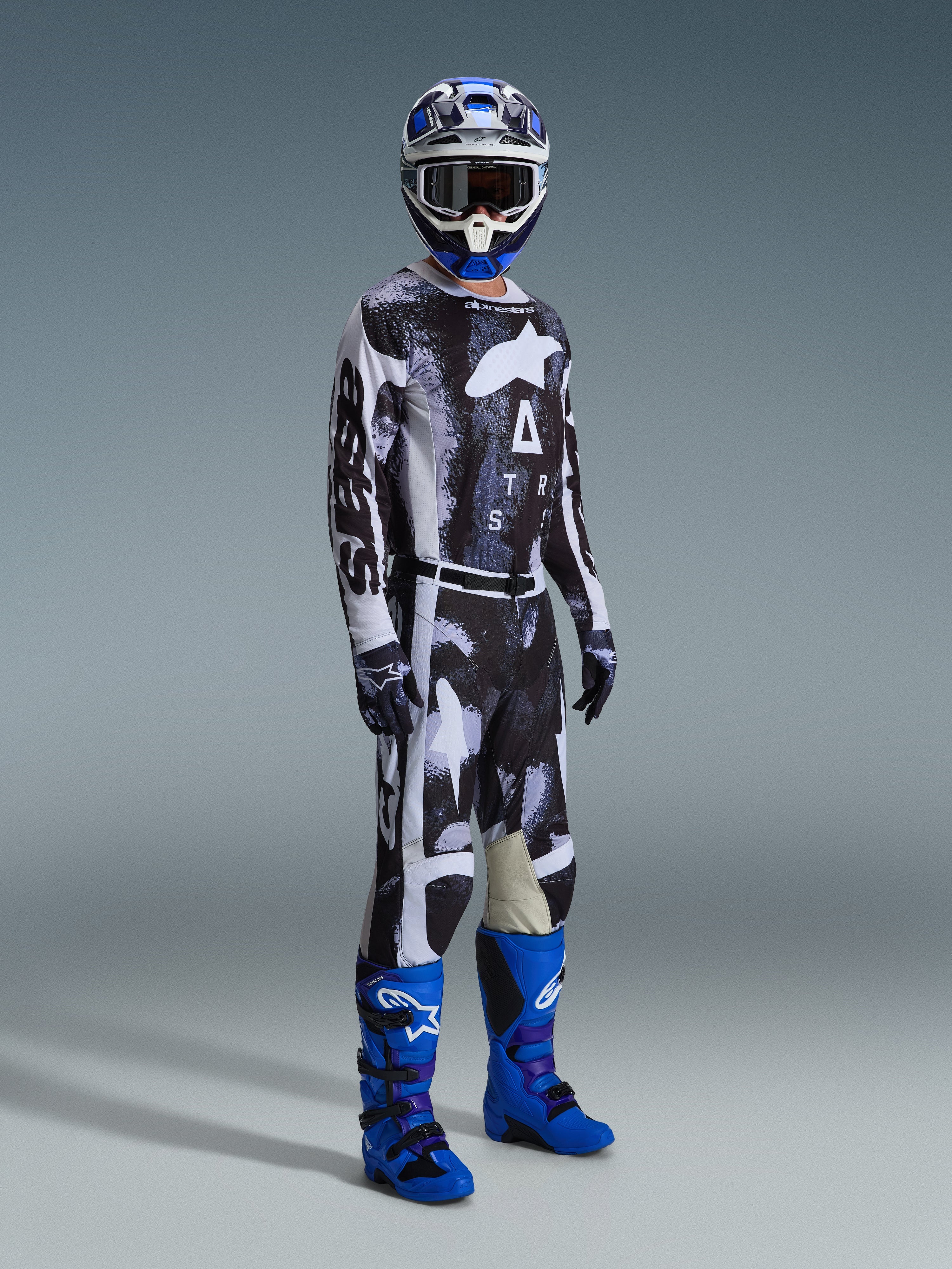 Motocross rider wearing the Alpinestars 2026 Racer Lahnd Jersey and matching pants in Iron Camo, featuring a black and white graphic pattern, paired with blue boots, a blue and white helmet, and black gloves, full racing gear for off-road riding.
